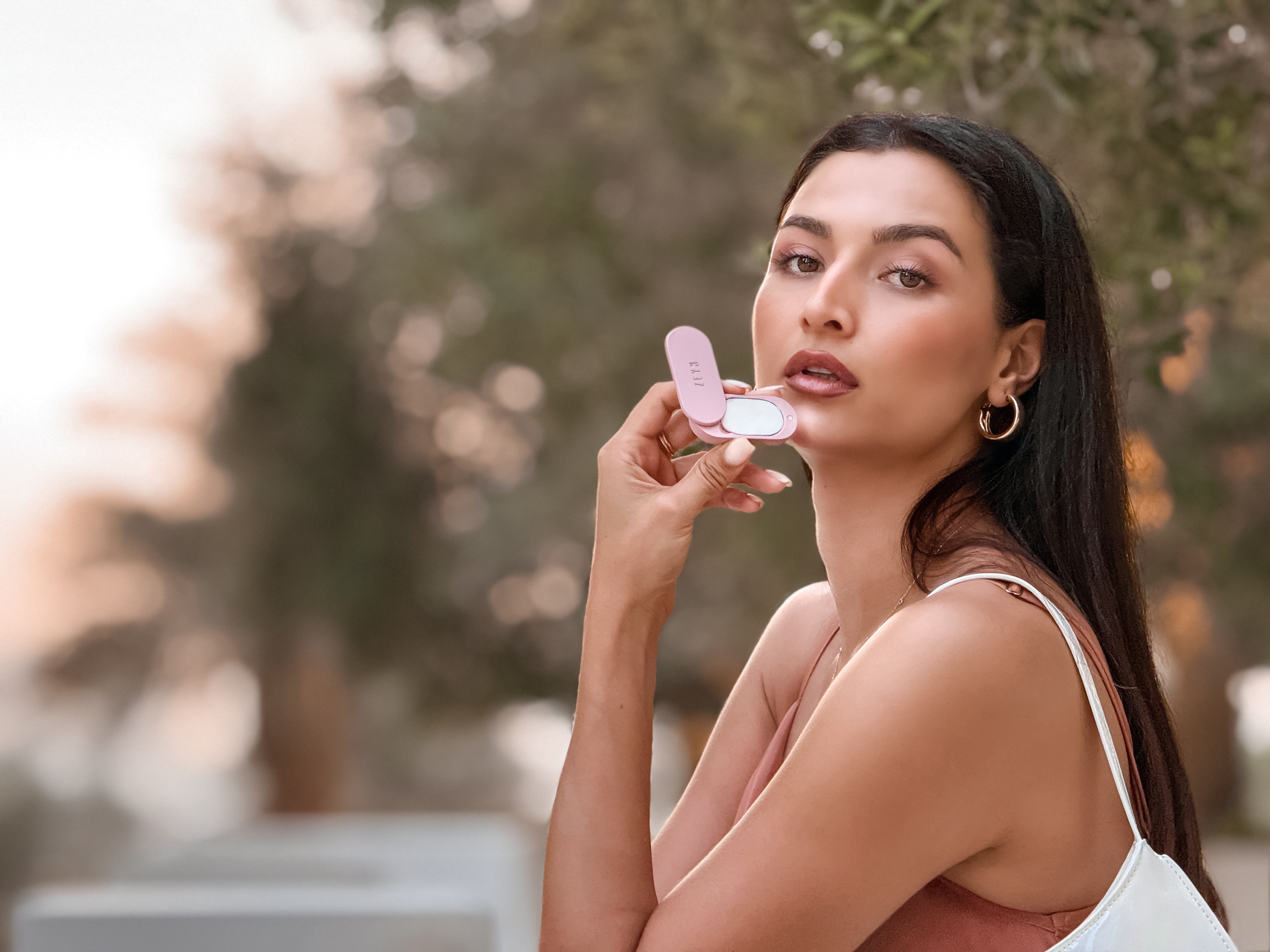 Woman holding a compact mirror outdoors with blurred trees in the background