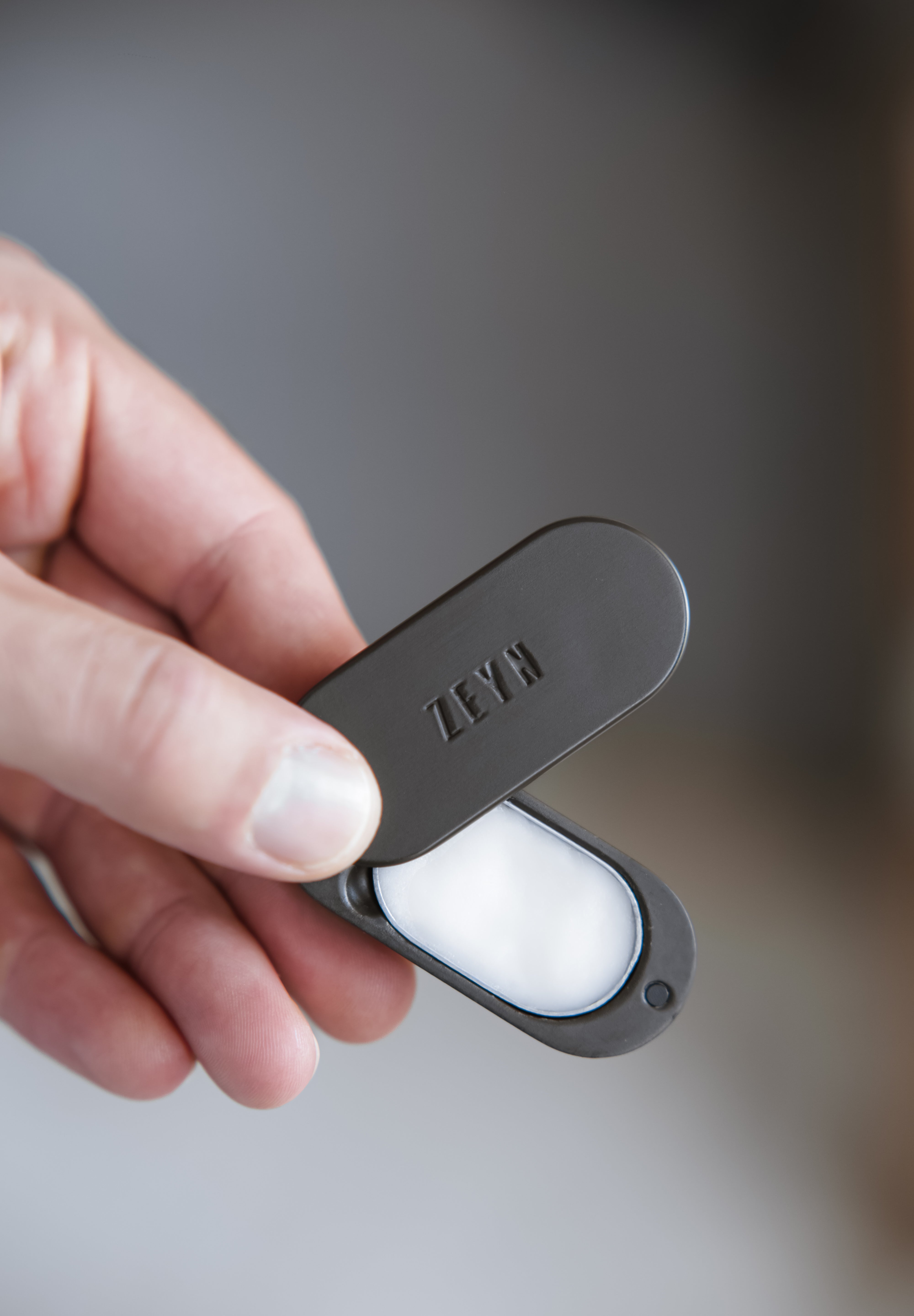 Hand holding a black case with 'Zeyn' branding, containing a white object on a blurred background.