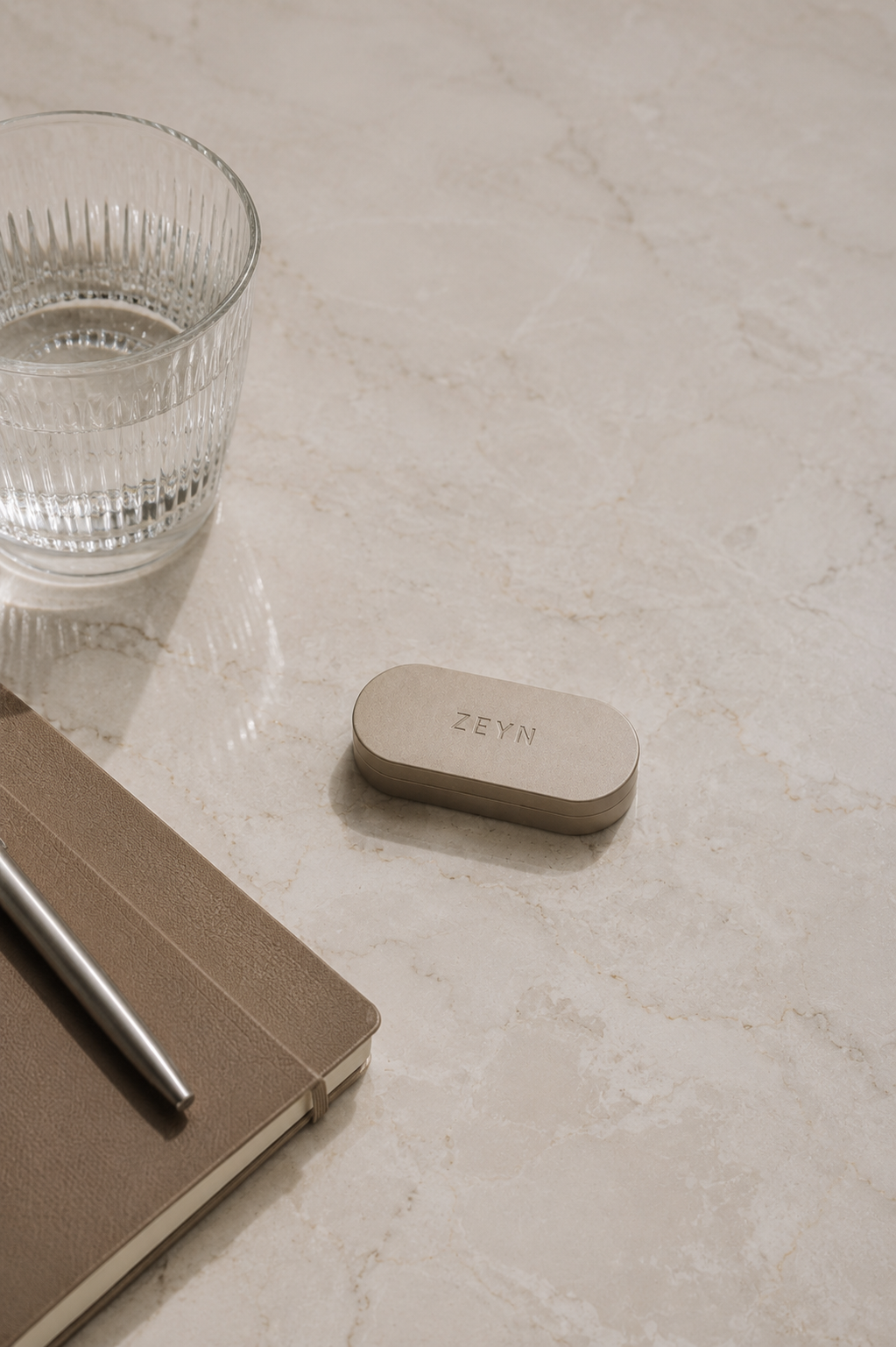 ZEYN warm stone refillable fragrance case on a light marble surface beside glassware in a refined everyday lifestyle setting